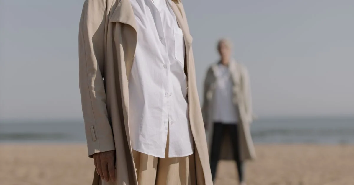 Mujeres de moda en abrigos beige están en una playa de arena, exhibiendo elegancia.