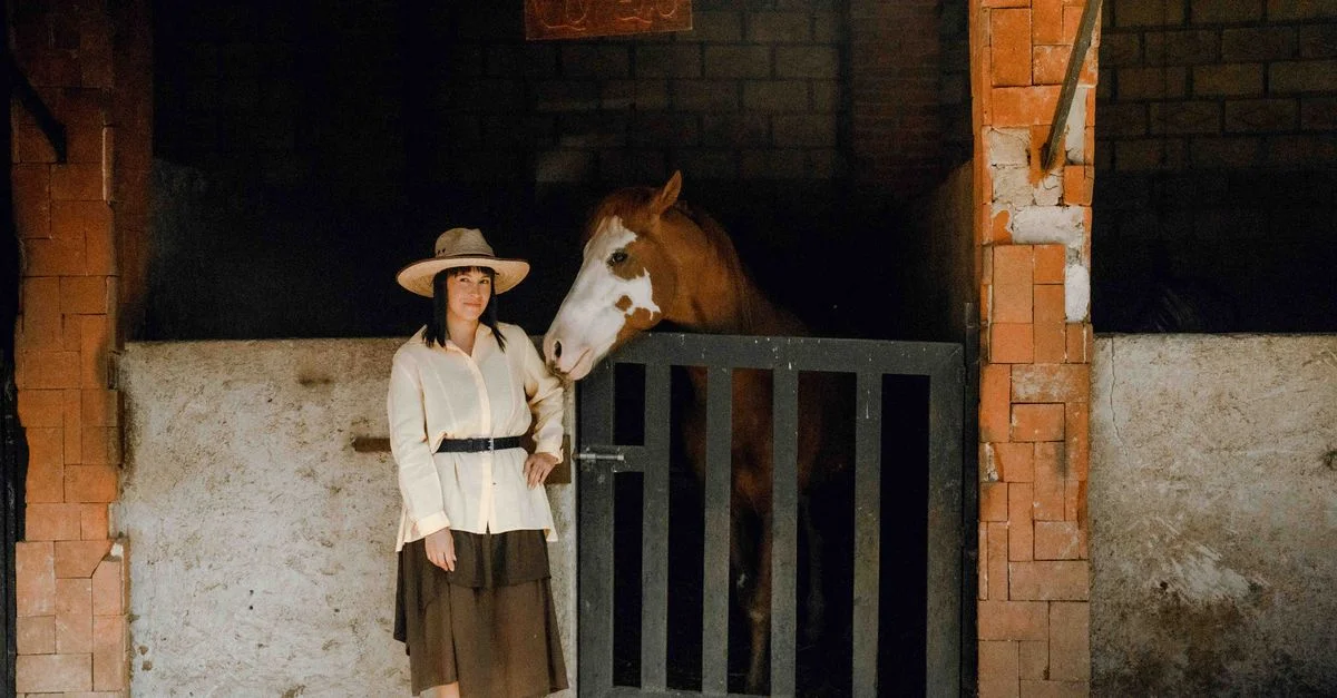 Una mujer vestida de vaquera junto a un caballo en un granero rústico.