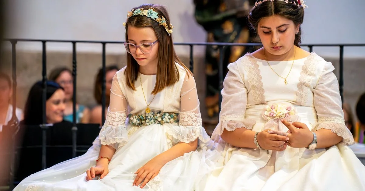 Dos chicas con vestidos blancos y diademas de flores se sientan en el interior durante una ceremonia, creando una atmósfera serena.